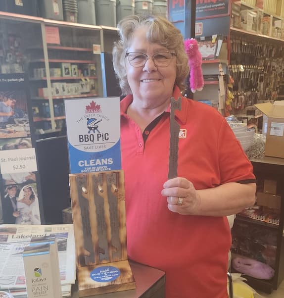 Photo of a Cashier at Home Hardware Retail store holding a BBQ Pic in her left hand and smiling. She's behind the The wooden BBQ Pic stand that has a few BBQ Pics hanging on its three wooden dowels. There's a card on the top of the display that has Made in Canada Logo on it with BBQ Pic logo in the middle and below it says "Easy to Use: Cleans Top of and bottom of grates".